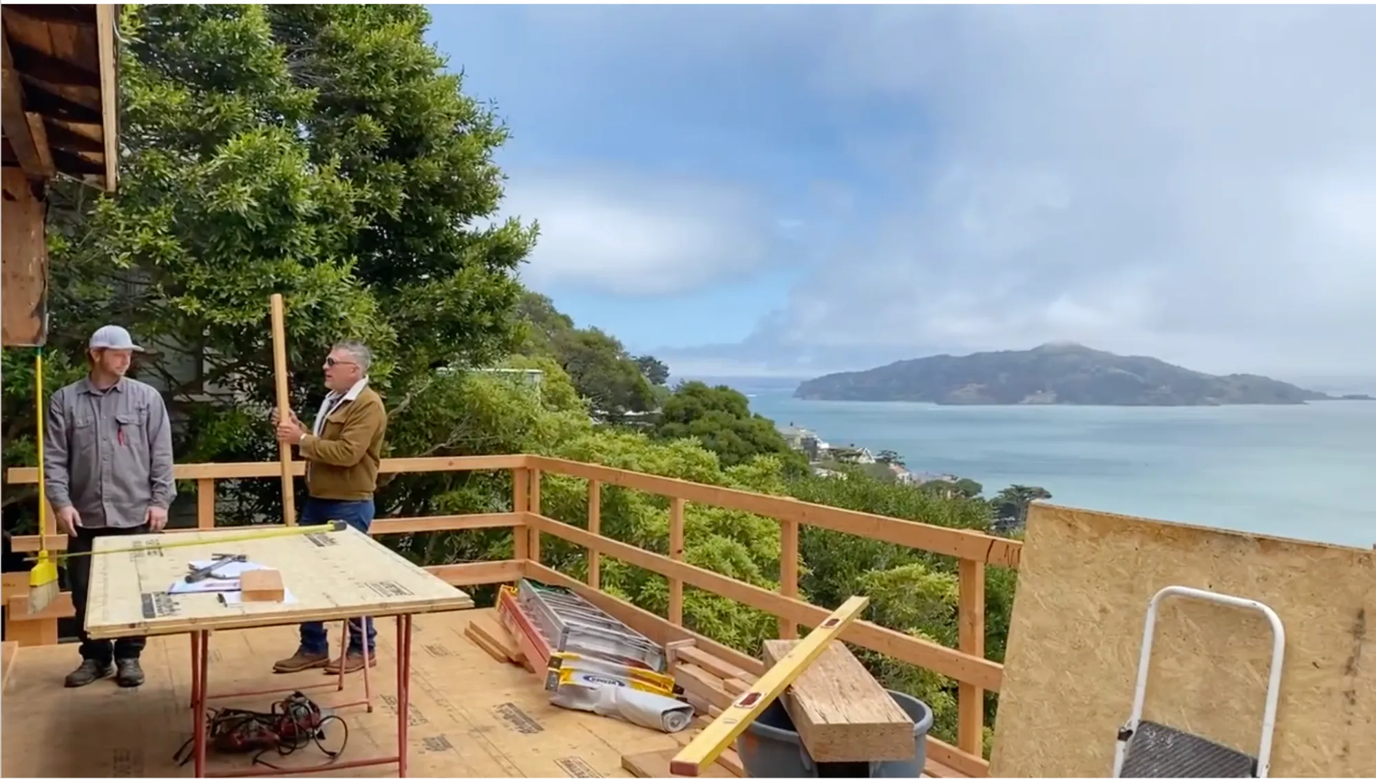 Two of the team at a worktable on the new deck, with timber framing behind them and Angel Island across the bay.