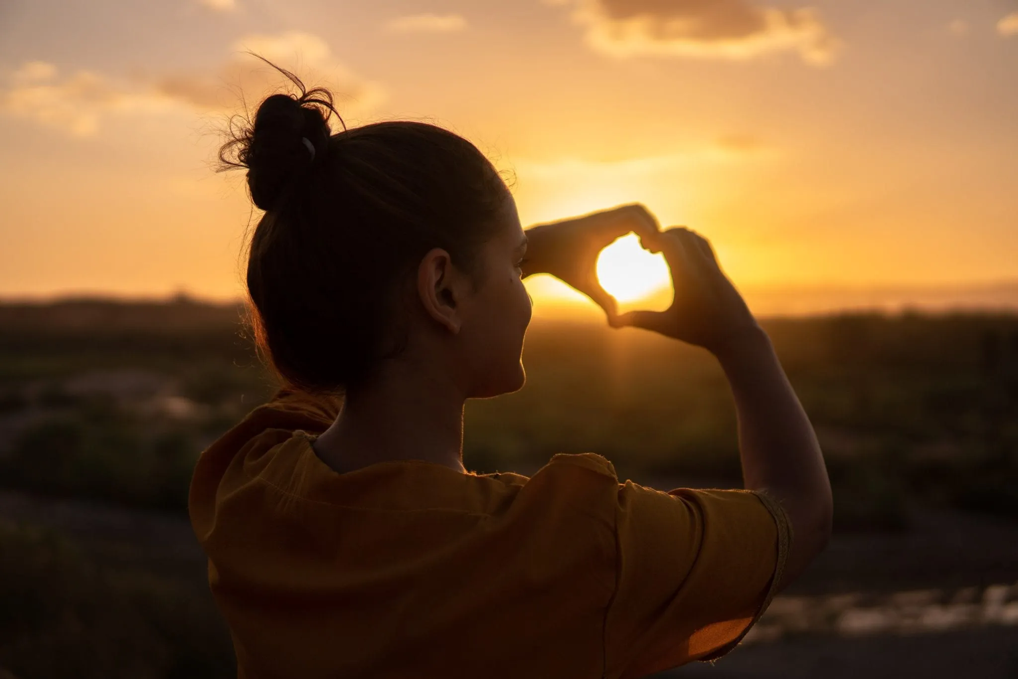 A woman forms a heart with her hands around the setting sun.