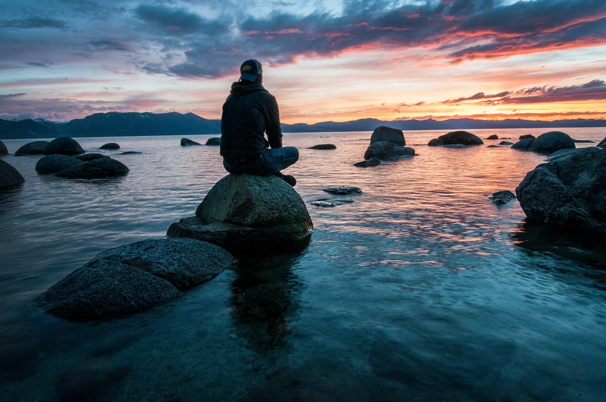 A person sits on a rock looking across a still ocean at sunset.